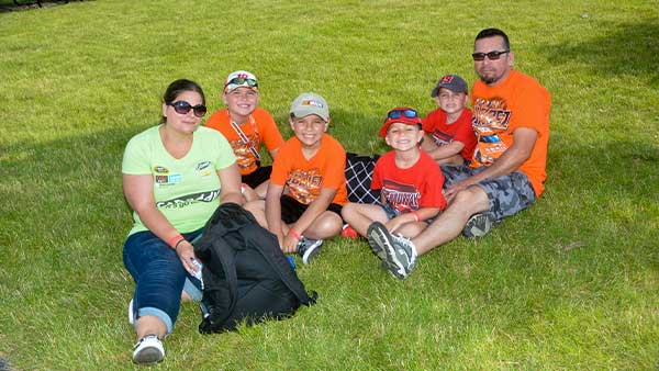 A family sitting together on green grass outdoors, smiling at the camera. Two adults and four children wear casual clothing, with several in bright orange shirts and baseball caps, suggesting attendance at a racing or sports event on a sunny day.